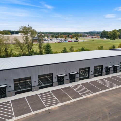 Aerial view of a long, gray industrial building with multiple glass garage-style doors and a striped parking lot.