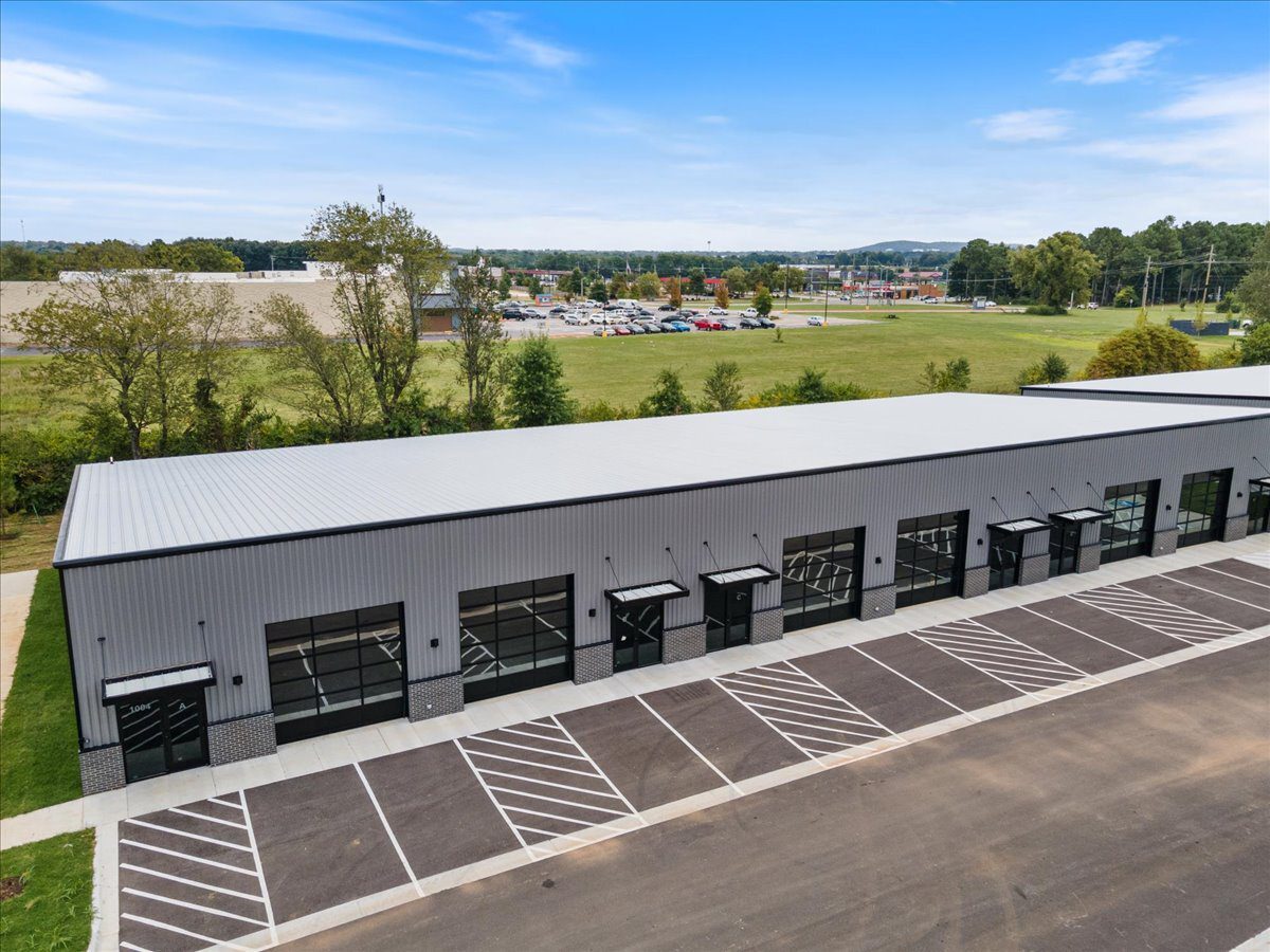 Aerial view of a long, gray industrial building with multiple glass garage-style doors and a striped parking lot.