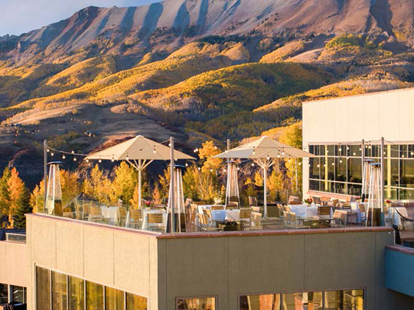 Rooftop terrace with patio tables, umbrellas, and string lights overlooking autumn-colored hills and mountains.